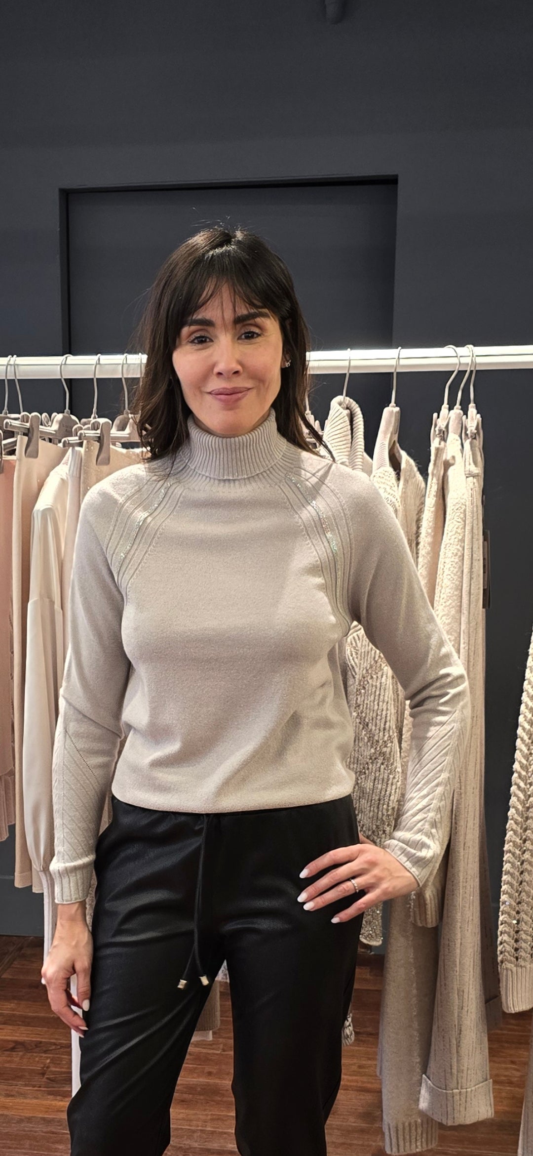 Woman wearing a light gray turtleneck sweater and black pants standing in front of a rack of clothes.